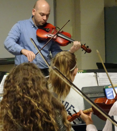 Teacher playing a violin as students watch and hold their own violins